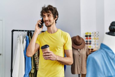 Young hispanic man talking on the smartphone drinking coffee at clothing store