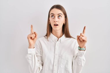 Young caucasian woman standing over isolated background amazed and surprised looking up and pointing with fingers and raised arms. 