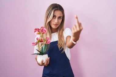 Young blonde woman wearing gardener apron holding plant showing middle finger, impolite and rude fuck off expression 