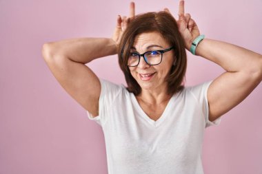 Orta yaşlı İspanyol bir kadın pembe arka planda durup komik ve çılgın pozlar veriyor parmakları kafasında tavşan kulağı, neşeli bir gülümseme 