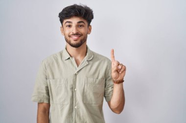 Arab man with beard standing over white background showing and pointing up with finger number one while smiling confident and happy. 