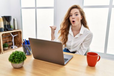 Young caucasian woman working at the office using computer laptop surprised pointing with hand finger to the side, open mouth amazed expression. 