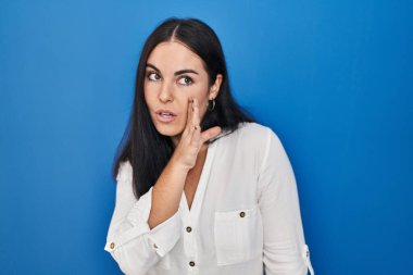 Latin kökenli genç bir kadın mavi arka planda el ele durur gizli dedikodular anlatır, kötü niyetli konuşmalar fısıldar. 