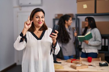 Three women working at small business ecommerce smiling with an idea or question pointing finger with happy face, number one 