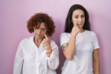 İspanyol anne ve kızı pembe arka plan üzerine gündelik beyaz tişört giyip senin jestini, şüpheli ifadesini izleyerek göze işaret ediyorlar. 