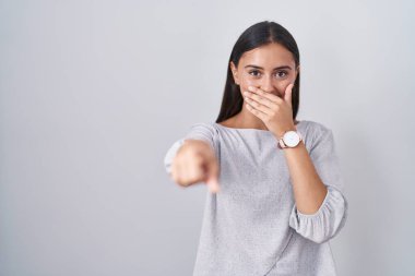 Young hispanic woman standing over white background laughing at you, pointing finger to the camera with hand over mouth, shame expression 