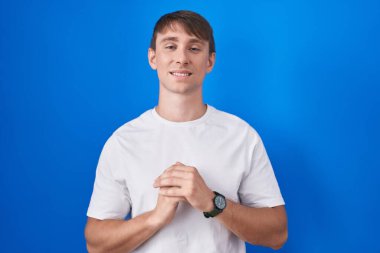 Caucasian blond man standing over blue background hands together and fingers crossed smiling relaxed and cheerful. success and optimistic 
