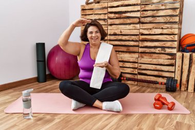 Middle age hispanic woman sitting on training mat at the gym gesturing with hands showing big and large size sign, measure symbol. smiling looking at the camera. measuring concept. 