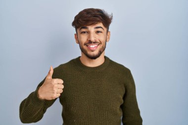 Arab man with beard standing over blue background doing happy thumbs up gesture with hand. approving expression looking at the camera showing success. 