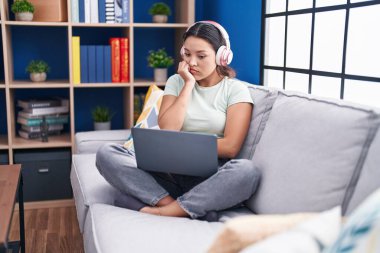 Hispanic young woman using laptop at home wearing headphones looking stressed and nervous with hands on mouth biting nails. anxiety problem. 