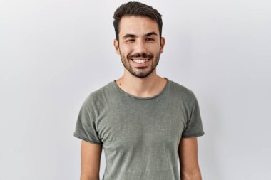 Young hispanic man with beard wearing casual t shirt over white background winking looking at the camera with sexy expression, cheerful and happy face. 