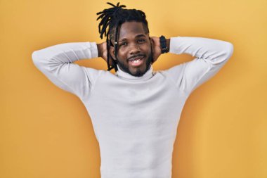 Sarı arka plan üzerine balıkçı yaka kazak giyen Afrikalı adam rahatlıyor ve esniyor, kolları ve elleri başının ve boynunun arkasında mutlu bir şekilde gülümsüyor. 