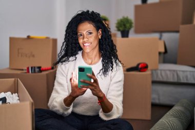 Afrika kökenli Amerikalı kadın akıllı telefon kullanıyor. Yeni evinde yerde oturuyor.