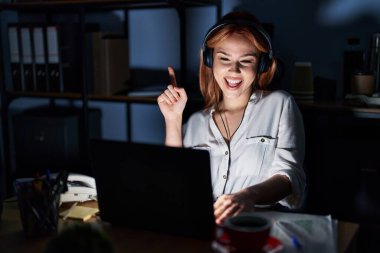 Geceleri ofiste çalışan beyaz bir kadın yüzünde kocaman bir gülümsemeyle kameraya el ve parmak ile bakıyor.. 