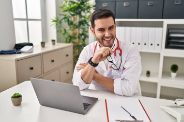 Klinikte çalışan dizüstü bilgisayar kullanan doktor üniforması giyen genç bir İspanyol.