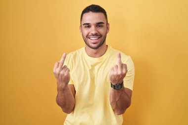 Young hispanic man standing over yellow background showing middle finger doing fuck you bad expression, provocation and rude attitude. screaming excited 