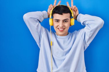 Non binary person listening to music using headphones posing funny and crazy with fingers on head as bunny ears, smiling cheerful 