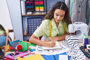 Young beautiful hispanic woman tailor smiling confident drawing on notebook at clothing factory