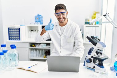 Laboratuvarda çalışan genç İspanyol adam laptopuyla mutlu ve olumlu gülümsüyor, başparmağıyla mükemmel ve onay işareti yapıyor. 