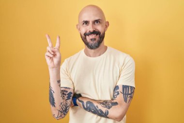 Sarı arka planda dikilen, gülümseyen, kameraya göz kırpan ve parmaklarıyla zafer işareti yapan İspanyol dövmeli adam. İki numara.. 