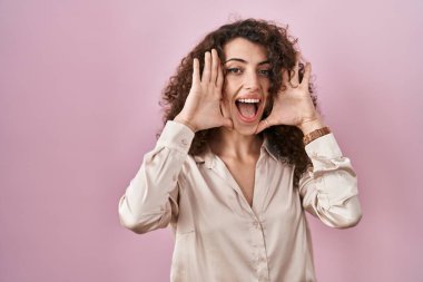 Hispanic woman with curly hair standing over pink background smiling cheerful playing peek a boo with hands showing face. surprised and exited 