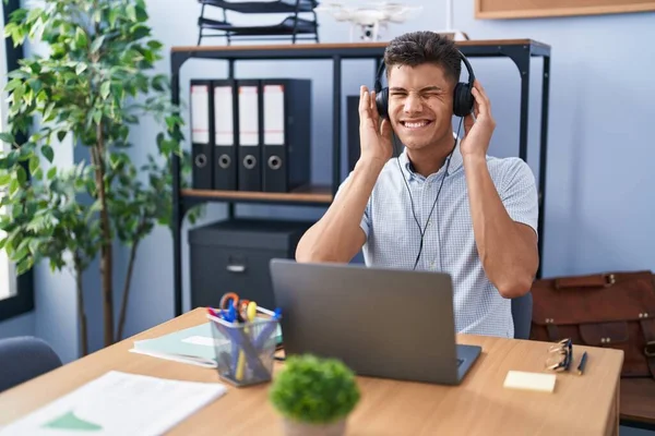 Young hispanic man working at the office wearing headphones covering ears with fingers with annoyed expression for the noise of loud music. deaf concept.
