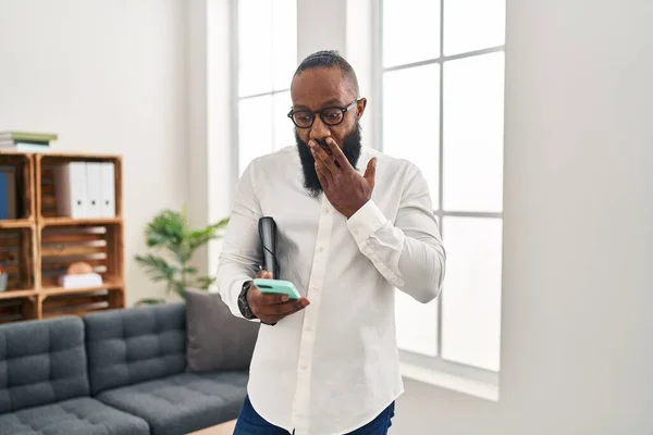 African american man working at therapy office using smartphone covering mouth with hand, shocked and afraid for mistake. surprised expression