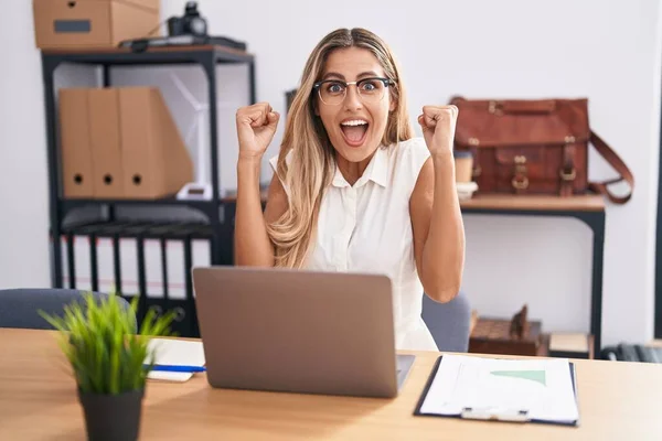 Young blonde woman working at the office wearing glasses screaming proud, celebrating victory and success very excited with raised arms