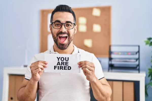 Young hispanic man with beard holding you are fired banner at the office smiling and laughing hard out loud because funny crazy joke.