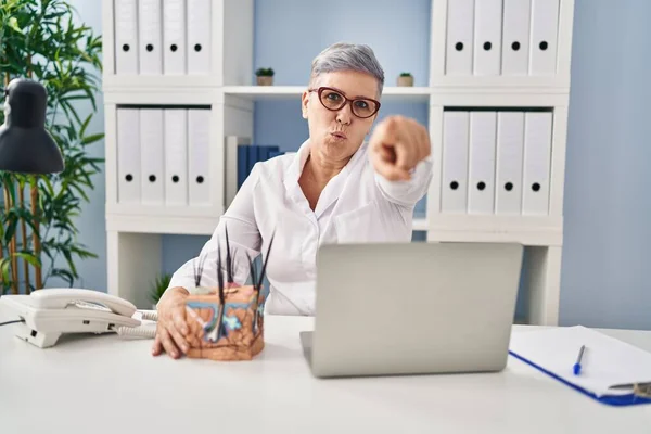 Middle age caucasian woman holding model of human anatomical skin and hair pointing with finger to the camera and to you, confident gesture looking serious