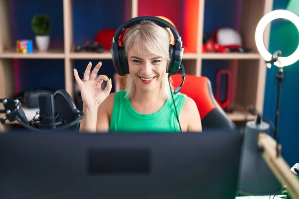 年轻的高加索女人带着耳机玩电子游戏 手拿着手签 笑容可亲地摆出一副极好的手势 — 图库照片
