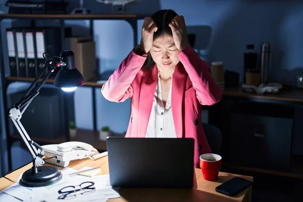 Chinese young woman working at the office at night suffering from headache desperate and stressed because pain and migraine. hands on head.