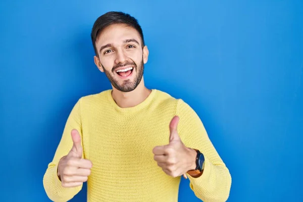 西班牙裔男子站在蓝色背景的成功标志上 用手做积极的手势 竖起大拇指 笑容满面 喜形于色 快乐的表达和胜利的姿态 — 图库照片