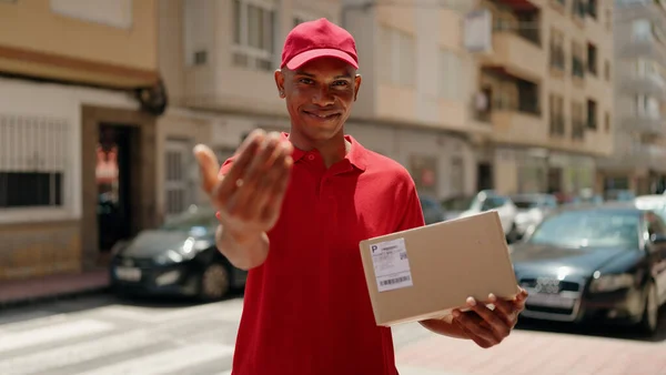 Young latin man delivery worker holding package doig coming gesture with hand at street