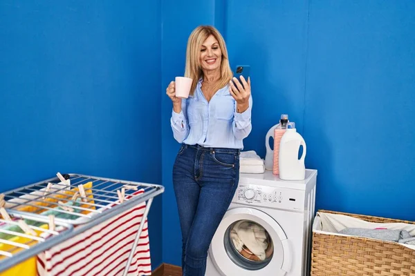 Middle age blonde woman using smartphone and drinking coffee waiting for washing machine at laundry room