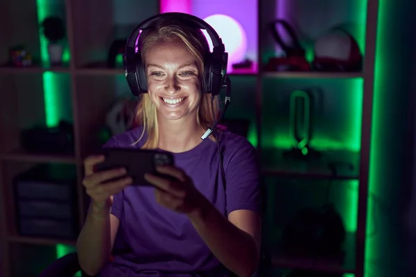 Young gamer woman playing video games with smartphone looking positive and happy standing and smiling with a confident smile showing teeth