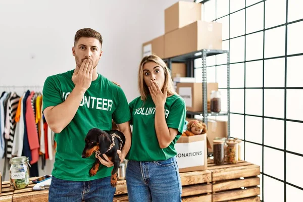 Young couple with cute dog wearing volunteer t shirt at donations stand covering mouth with hand, shocked and afraid for mistake. surprised expression