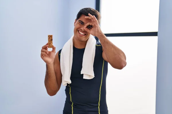 Hispanic man eating protein bar as healthy energy snack smiling happy doing ok sign with hand on eye looking through fingers