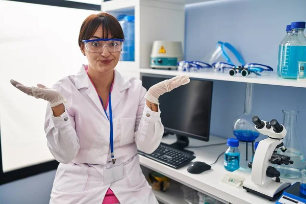 Young brunette woman working at scientist laboratory clueless and confused expression with arms and hands raised. doubt concept.