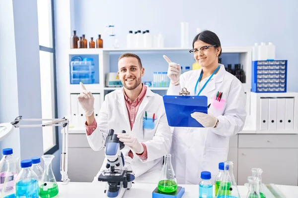Two young people working at scientist laboratory smiling happy pointing with hand and finger to the side