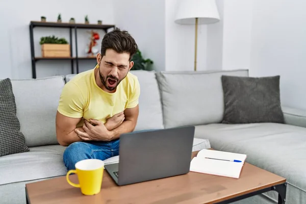 Young man with beard using laptop at home with hand on stomach because nausea, painful disease feeling unwell. ache concept.