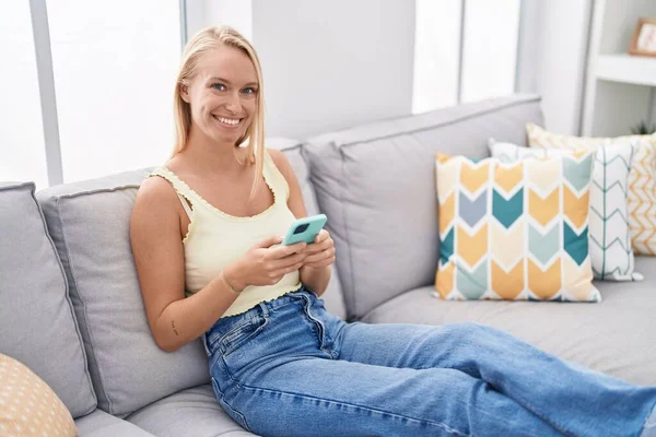 若いですブロンド女性使用してスマートフォン座っています上のソファにホーム — ストック写真