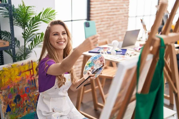 年轻的金发女艺术家微笑着自信地在艺术工作室的智能手机前自拍 — 图库照片