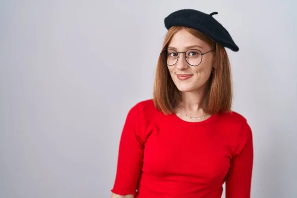 Young redhead woman standing wearing glasses and beret smiling looking to the side and staring away thinking.