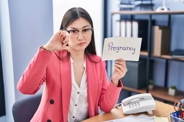 Chinese young woman working at the office holding pregnancy sign with angry face, negative sign showing dislike with thumbs down, rejection concept