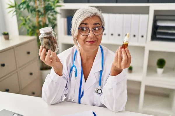 Middle age woman doctor holding cbd oil smiling looking to the side and staring away thinking.