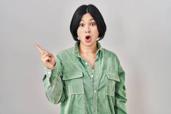Young asian woman standing over white background surprised pointing with finger to the side, open mouth amazed expression.