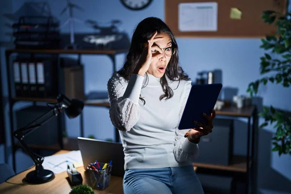 Young brazilian woman using touchpad at night working at the office doing ok gesture shocked with surprised face, eye looking through fingers. unbelieving expression.