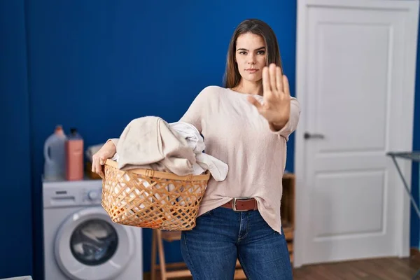 Beautiful woman holding laundry basket with open hand doing stop sign with serious and confident expression, defense gesture