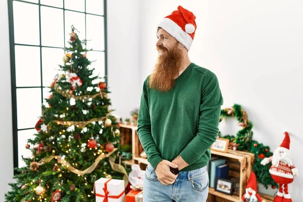 Redhead man with long beard wearing christmas hat by christmas tree looking away to side with smile on face, natural expression. laughing confident.